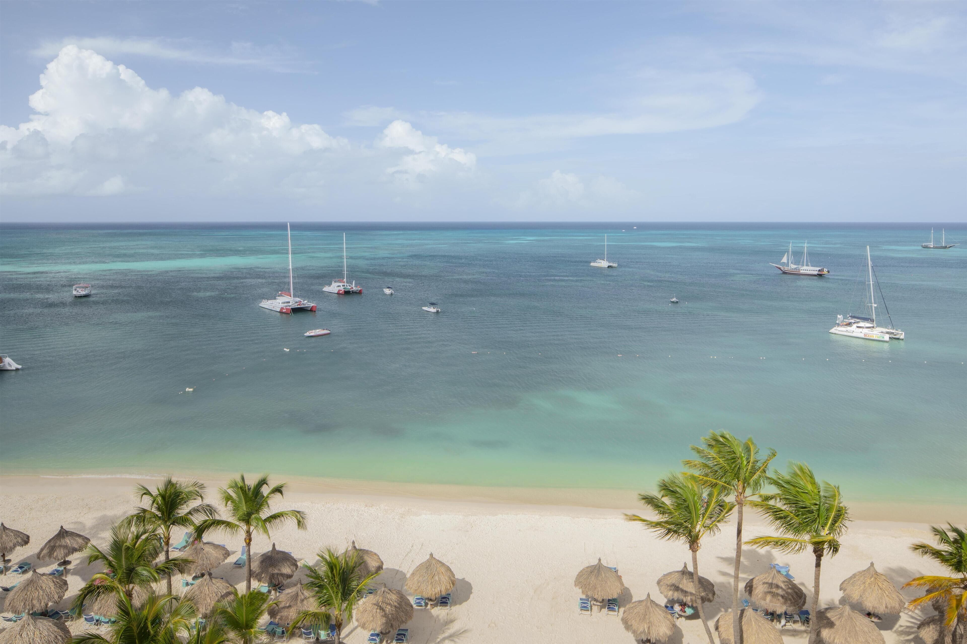 Una playa con sombrillas de paja, palmeras y barcos en el agua.