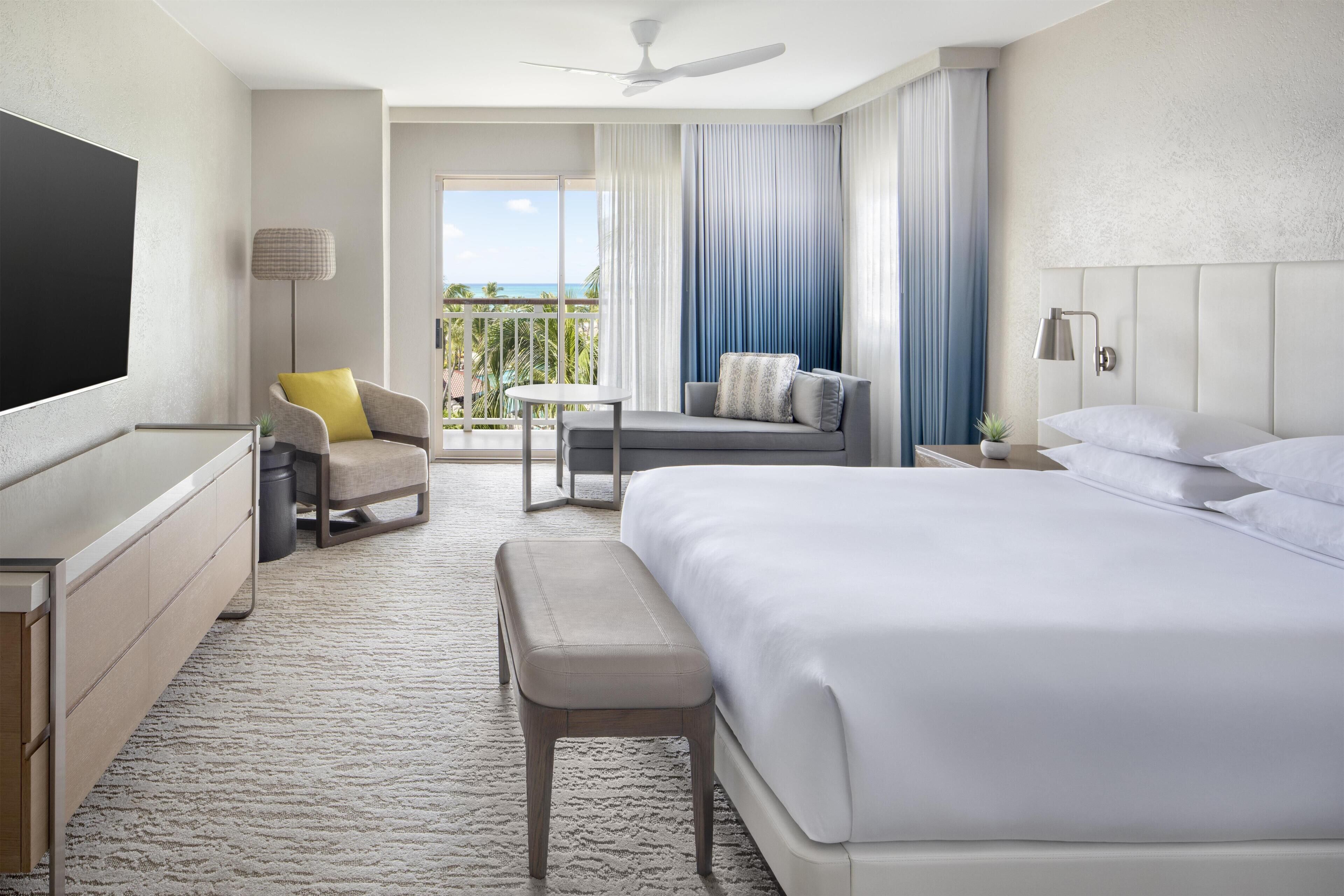 Habitación de hotel con cama, escritorio, silla, banco, televisor y vistas al exterior.