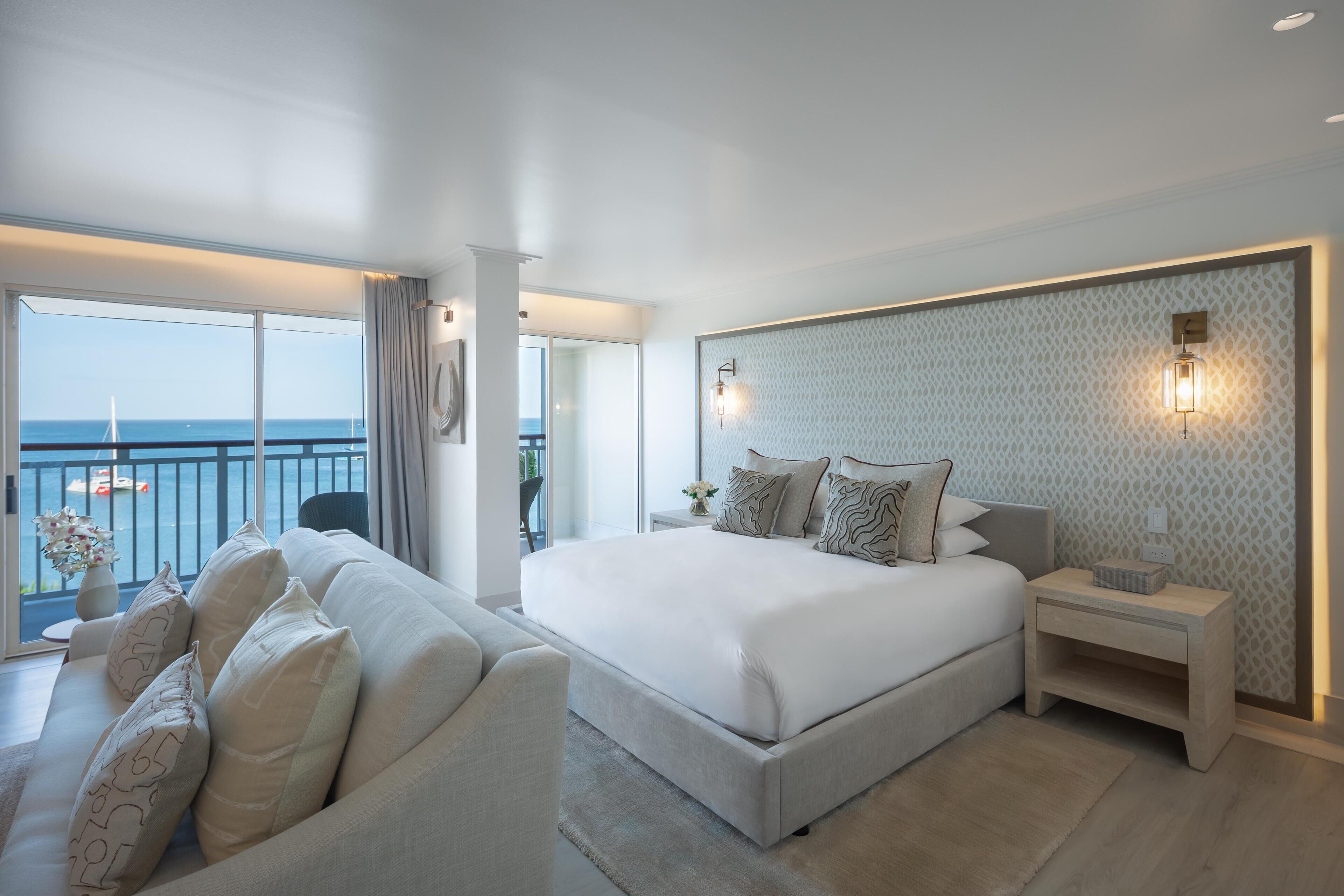 A modern hotel room with a large bed, a sofa, a nightstand, and a view of the sea.