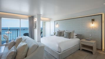 A modern hotel room with a large bed, a sofa, a nightstand, and a view of the sea.