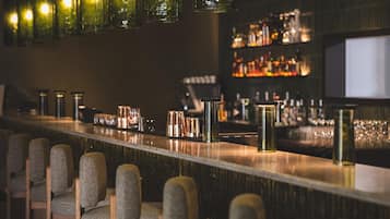 Un espace bar doté d’un long comptoir, de tabourets de bar et d’une étagère où sont disposées des bouteilles et des verres.