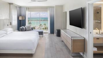 A modern hotel room with a large bed, a flat-screen TV, a balcony with a view of the ocean, and a bathroom with a sink and mirror.