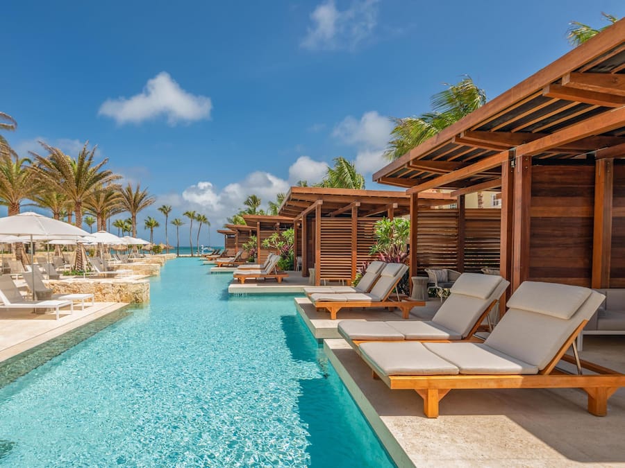 A poolside area with lounge chairs and umbrellas, surrounded by palm trees and a clear sky.