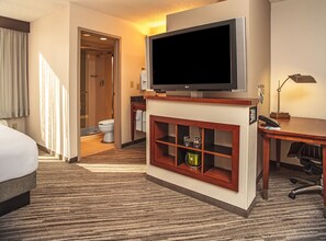Habitación de hotel con televisor de pantalla plana grande, un mueble de madera con un adorno, un escritorio con lámpara, un baño con ducha y una cama con cabecero.