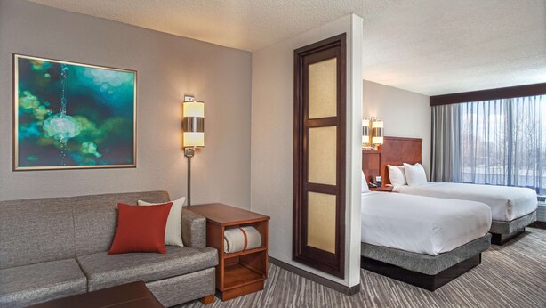 A hotel room with a grey sofa, a wooden side table, two beds with white linens, a large mirror, and a painting on the wall. - Hyatt Place Cincinnati/Blue Ash (Cincinnati)