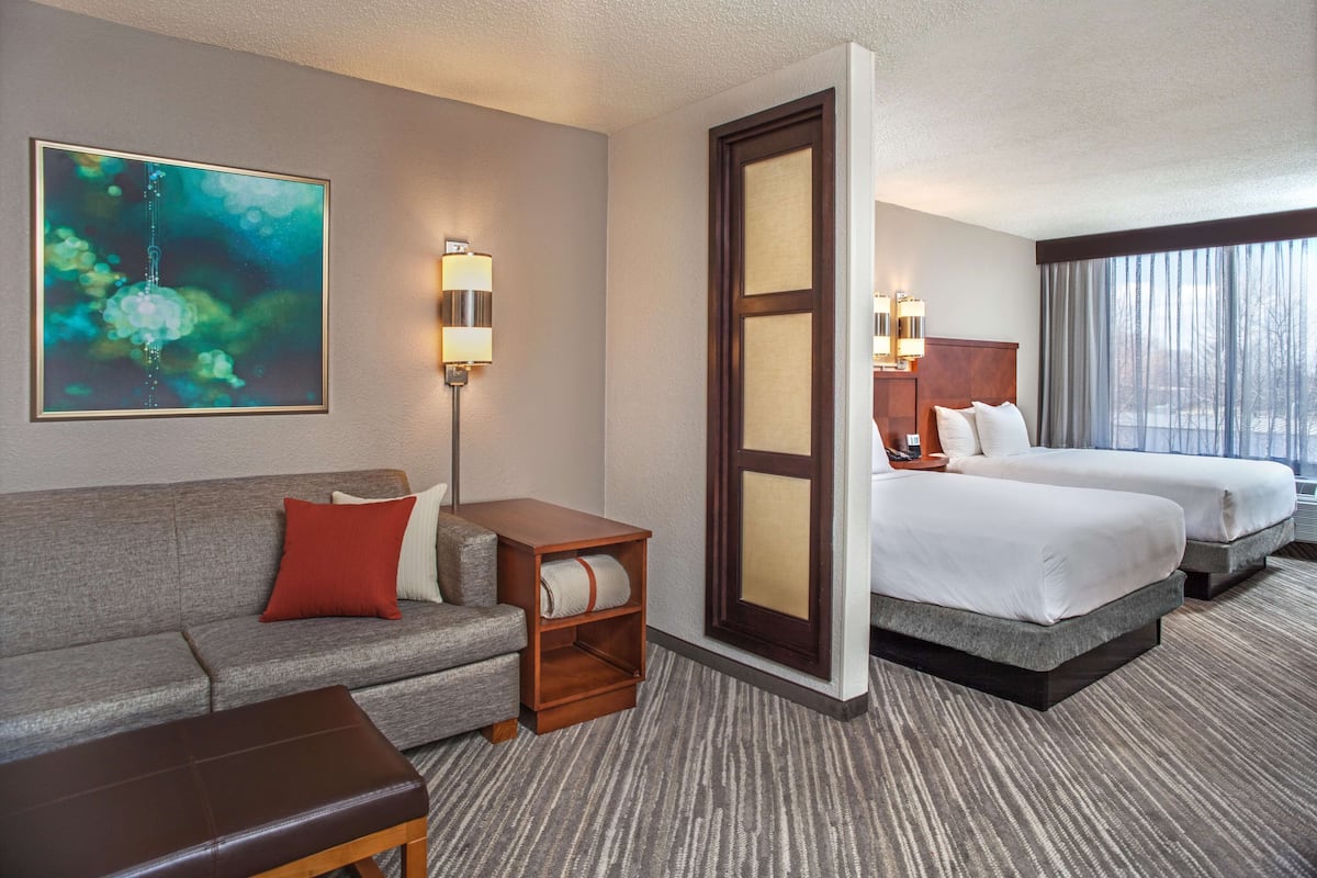 A hotel room with a grey sofa, a wooden side table, two beds with white linens, a large mirror, and a painting on the wall.