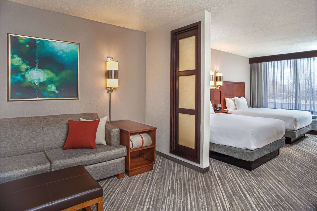 A hotel room with a sofa, a small table, a bedside table, two beds, a large window, and a painting on the wall.