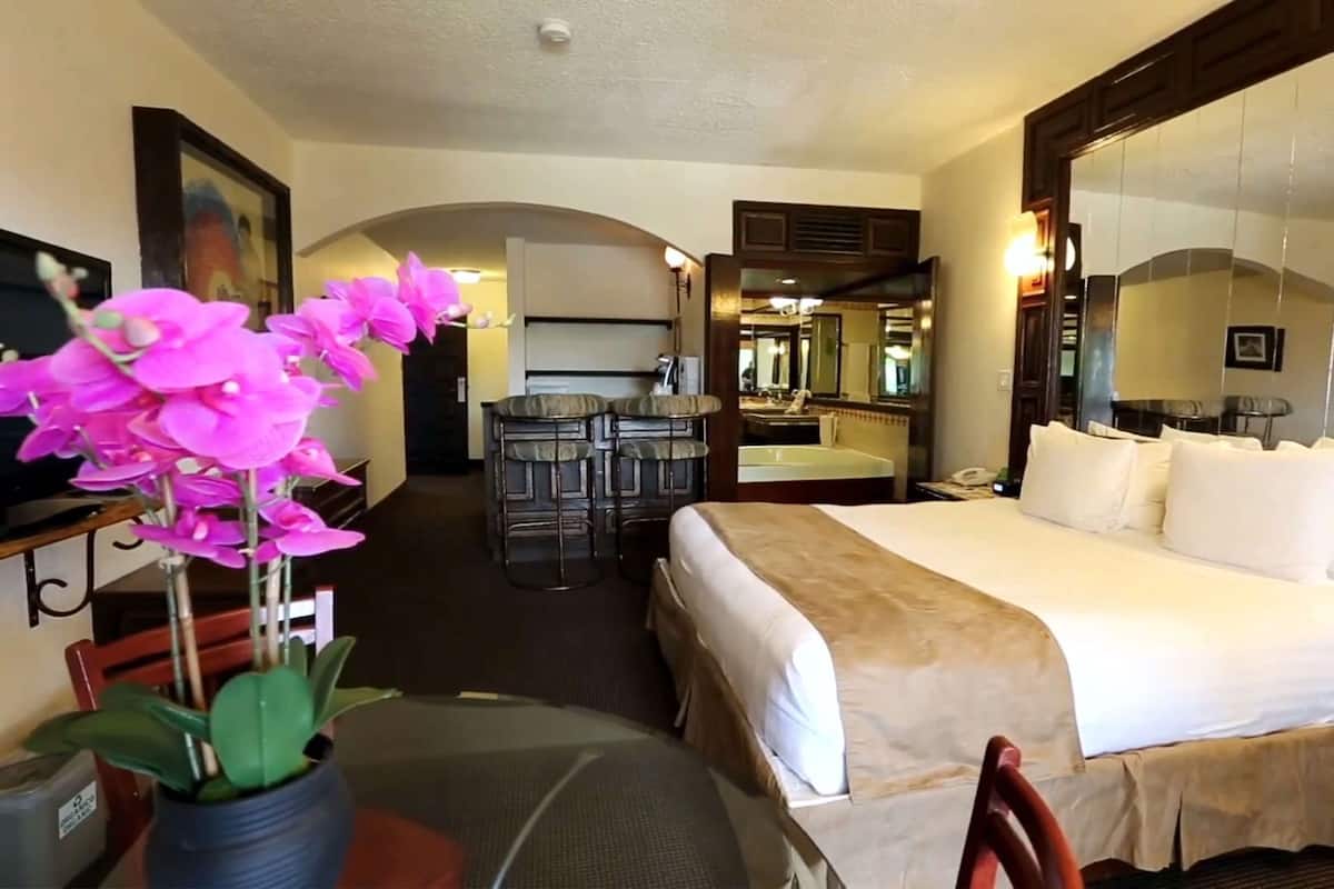 A hotel room with a bed, a desk, a chair, a small table, a mirror, and a potted plant.