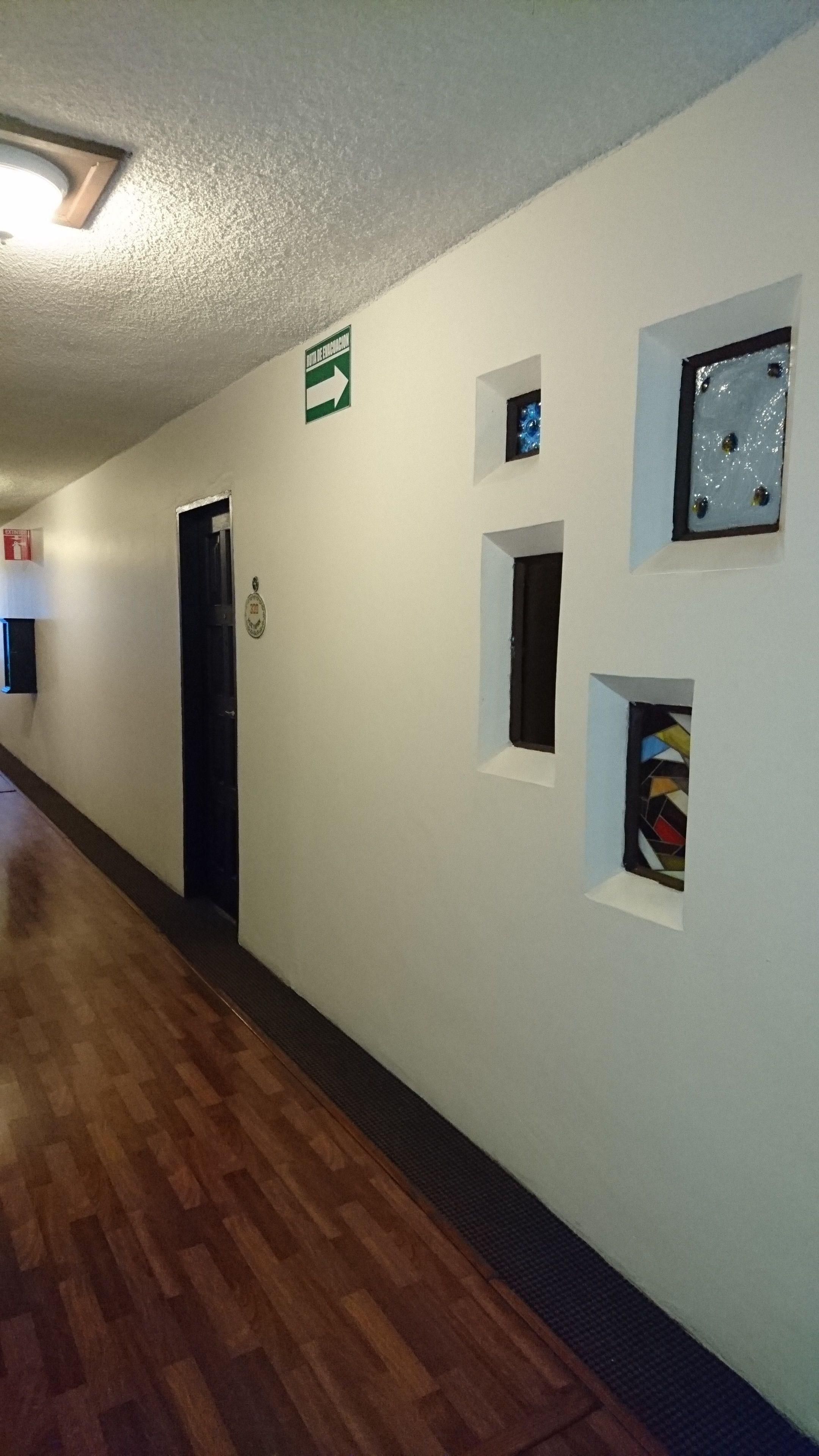 A hallway with a wooden floor, white walls, and several small windows. There is a green directional sign on the wall and a door with a plaque.