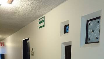 A hallway with a wooden floor, white walls, and several small windows. There is a green directional sign on the wall and a door with a plaque.