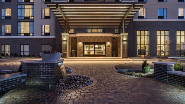 A hotel entrance with a covered walkway, a landscaped area with a fire feature, and a brick facade. - Hampton Inn & Suites Newport/Cincinnati (Newport)
