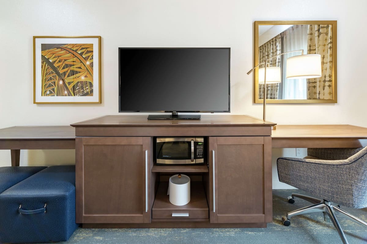 A hotel room with a flat-screen TV, a microwave, a small trash bin, a blue suitcase, and a chair.