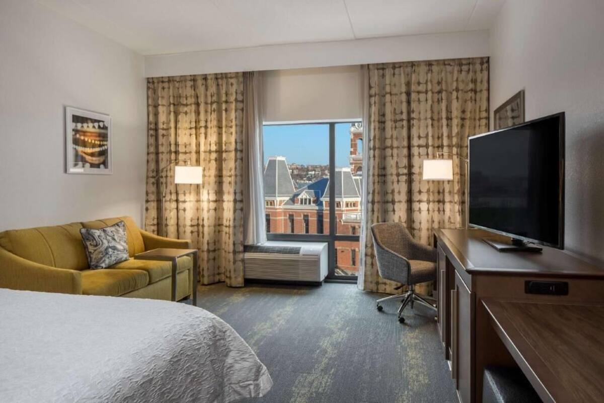 A hotel room with a bed, a sofa, a desk with a TV, a chair, and a large window with curtains.