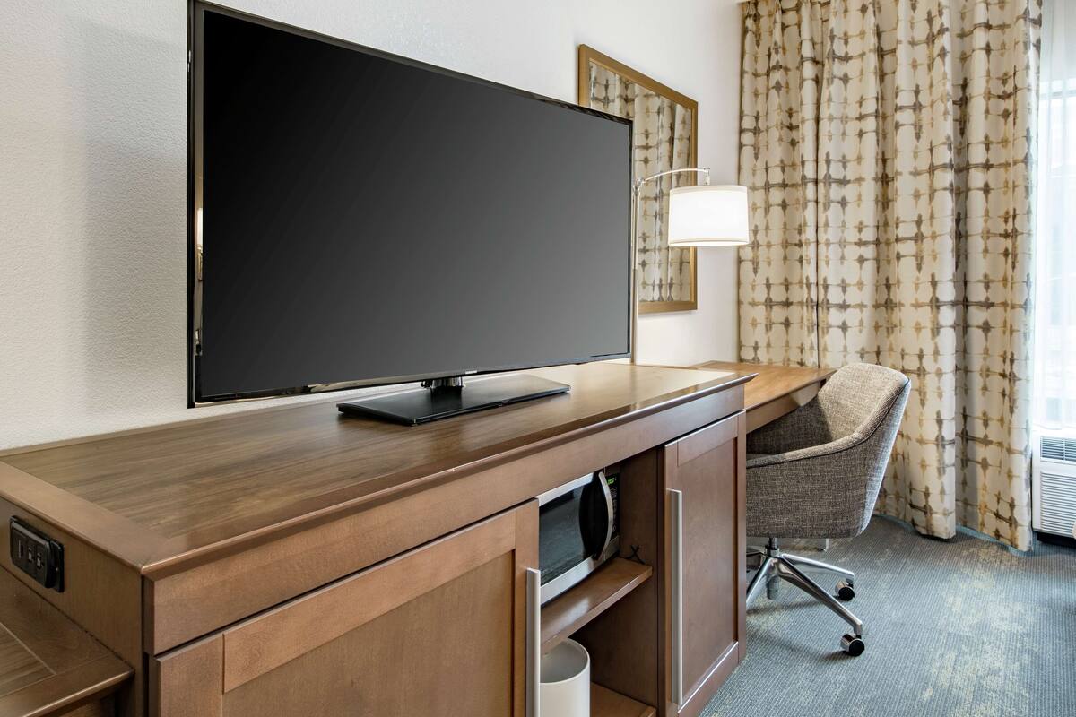 A hotel room with a wooden desk, a flat-screen TV, a chair, a lamp, and a patterned curtain.