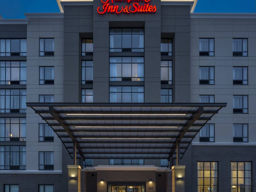 Un moderno edificio de hotel con un letrero destacado que dice “Hampton Inn & Suites”. Cuenta con una entrada cubierta con toldo de vidrio, varios pisos con amplias ventanas y fachada de ladrillo.