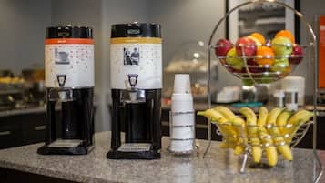 Dos dispensadores de café, una pila de tazas blancas y una cesta de fruta sobre una encimera.