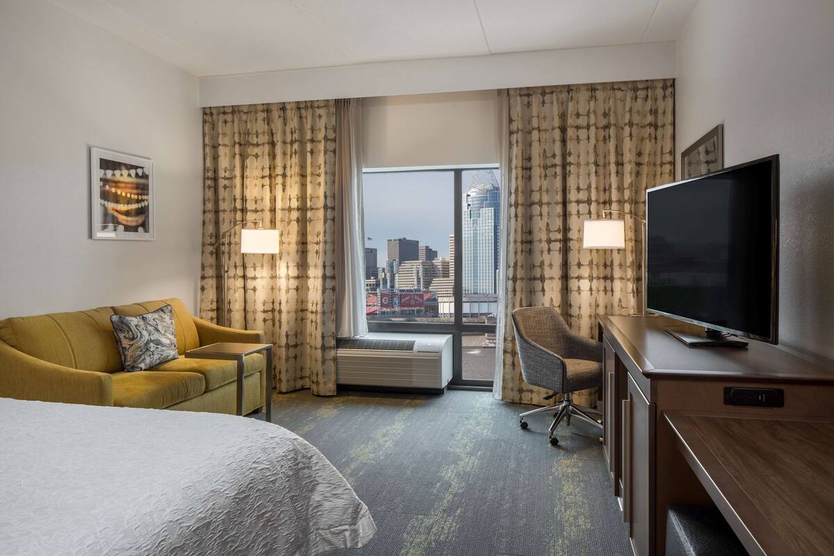 A hotel room with a bed, a sofa, a desk with a TV, a chair, and a large window with a city view.