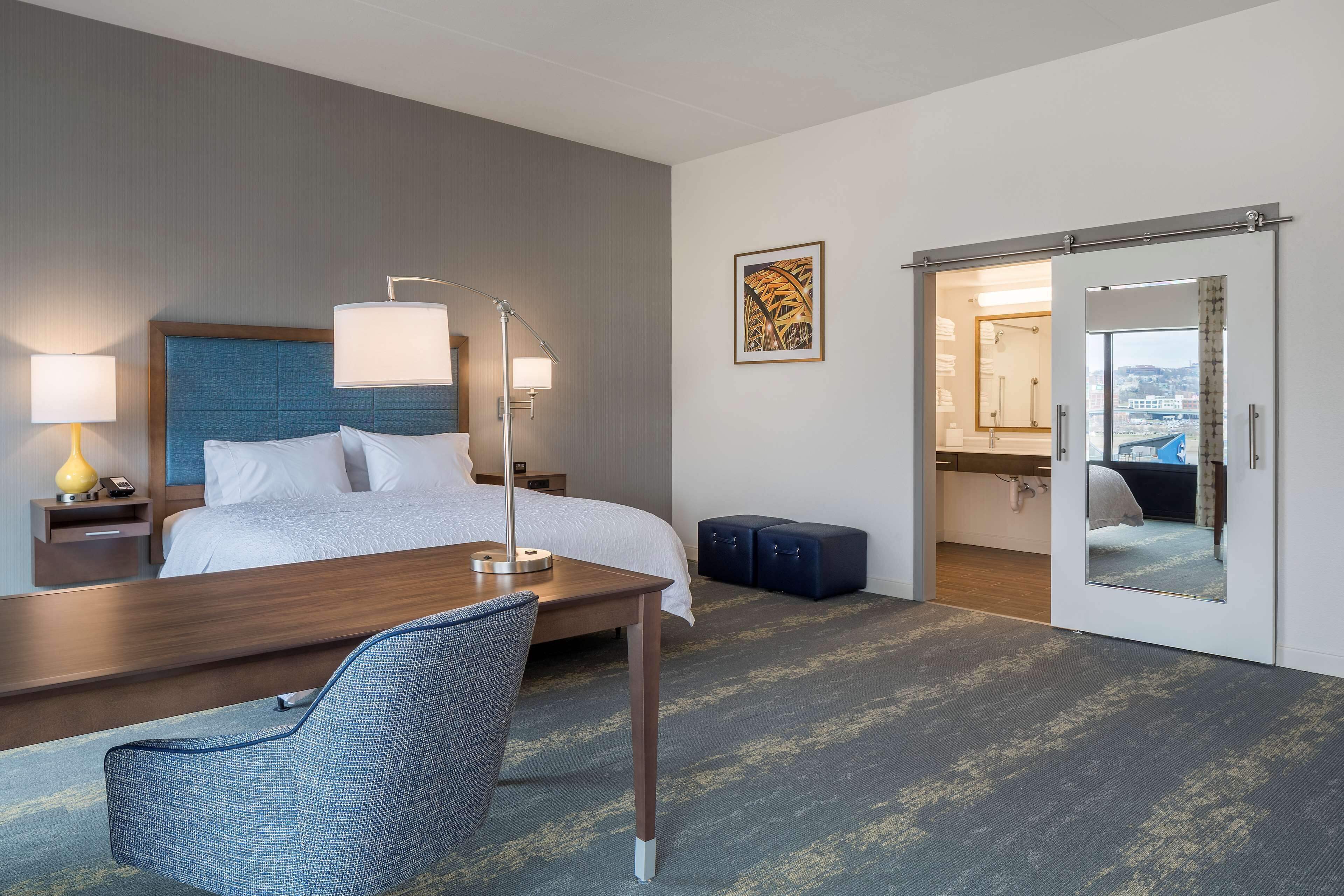 A hotel room with a bed, bedside lamps, a desk, a chair, a mirror, and a view of the city through the door.