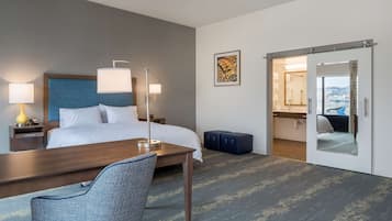 A hotel room with a bed, bedside lamps, a desk, a chair, a mirror, and a view of the city through the door.