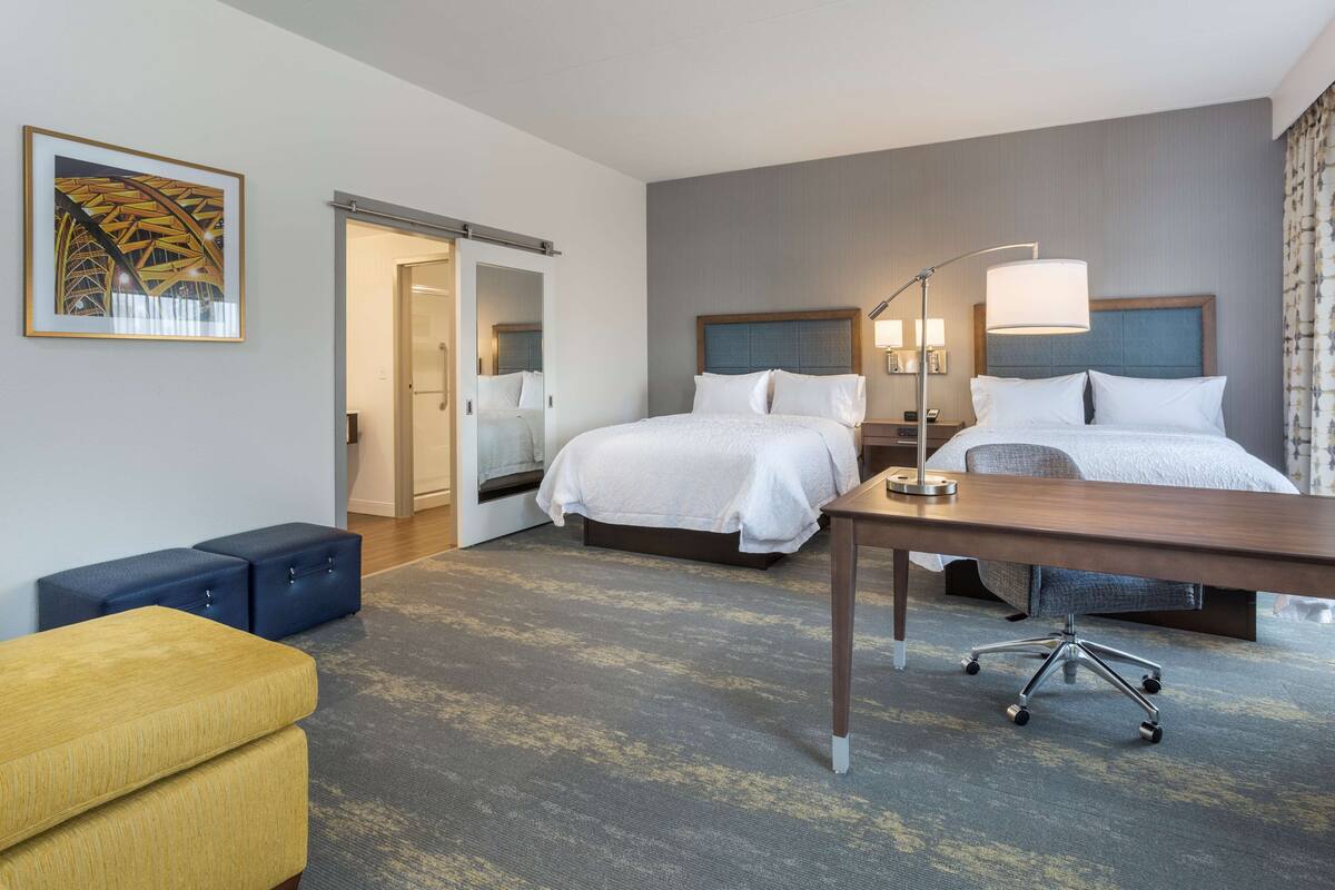 A hotel room with two beds, a desk, a chair, a sofa, and a painting on the wall.