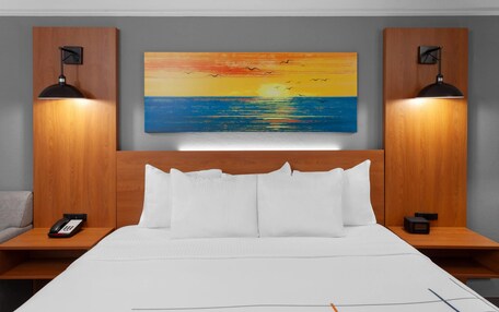 A hotel room with a large bed, two bedside tables, a painting of a sunset over the ocean, and two wall-mounted lamps.