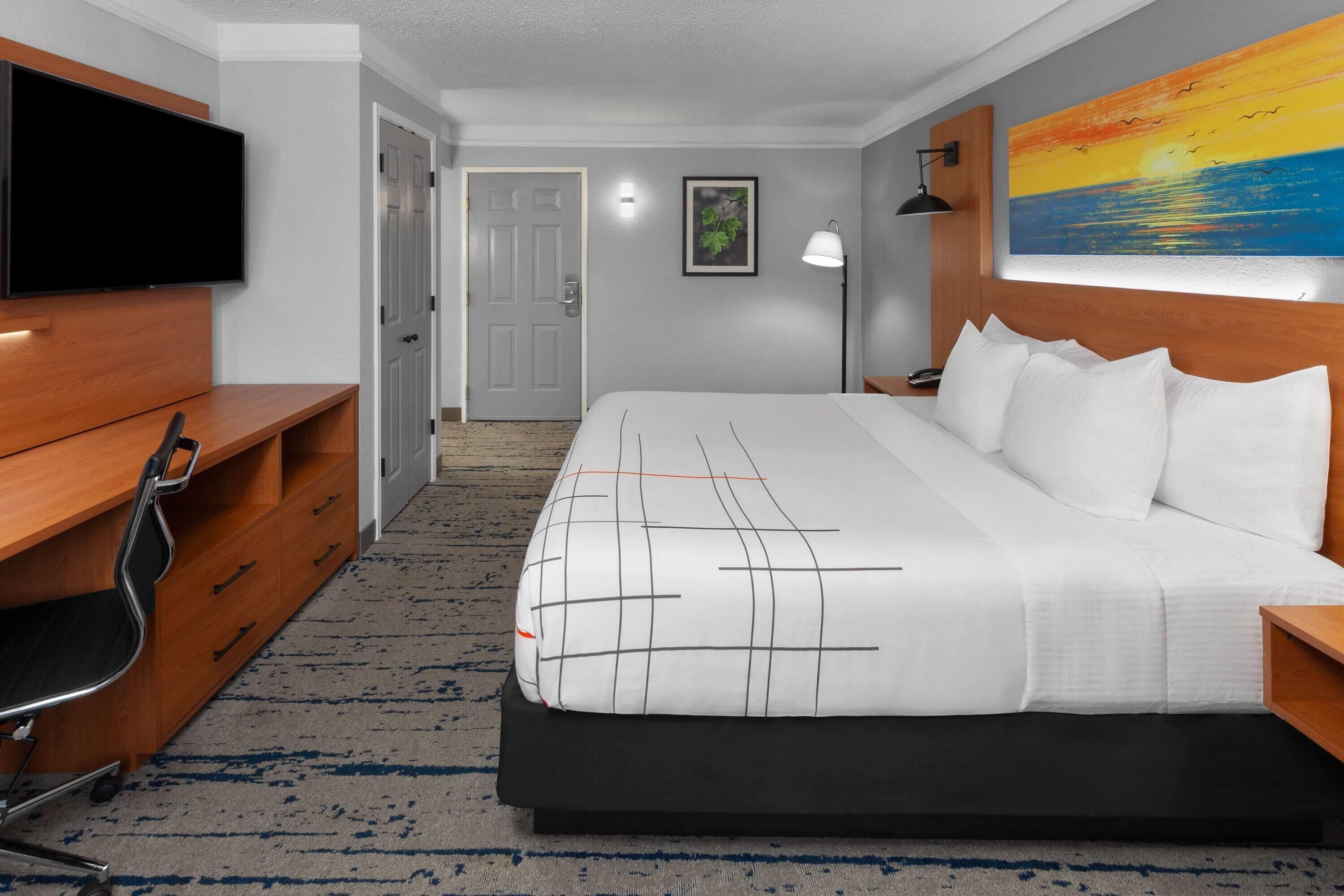 Habitación de hotel con una cama grande, un escritorio con silla, televisor de pantalla plana, un cuadro en la pared y una puerta que da a otra habitación.