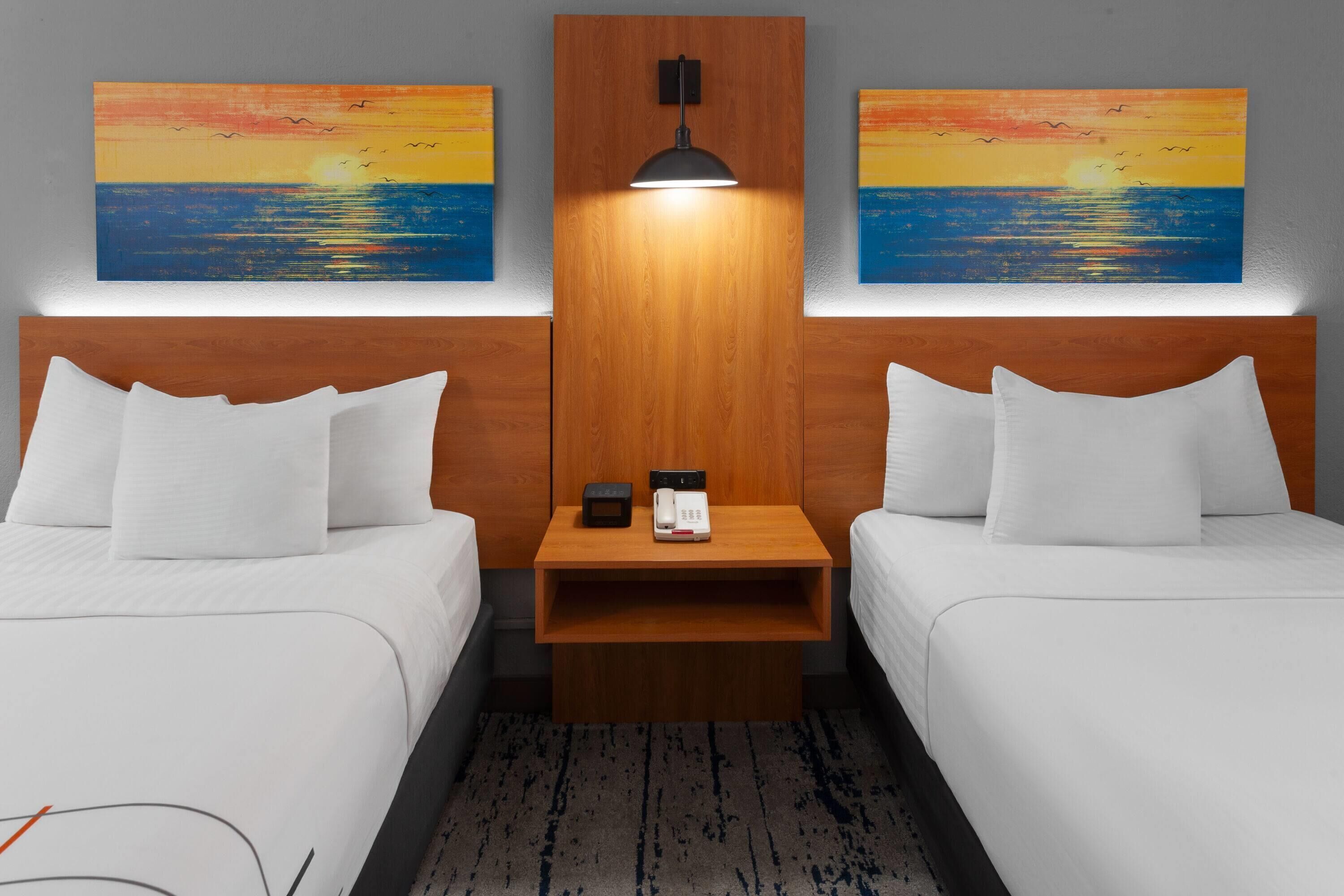 Een hotelkamer met twee bedden, een nachtkastje met een lamp, en twee schilderijen van een zonsondergang boven de oceaan aan de muur.