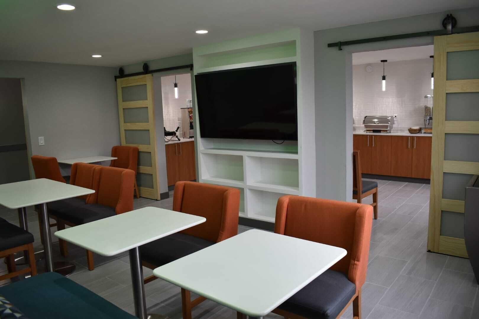 A modern dining area with tables and orange chairs, a large flat-screen TV, and a kitchen area in the background.