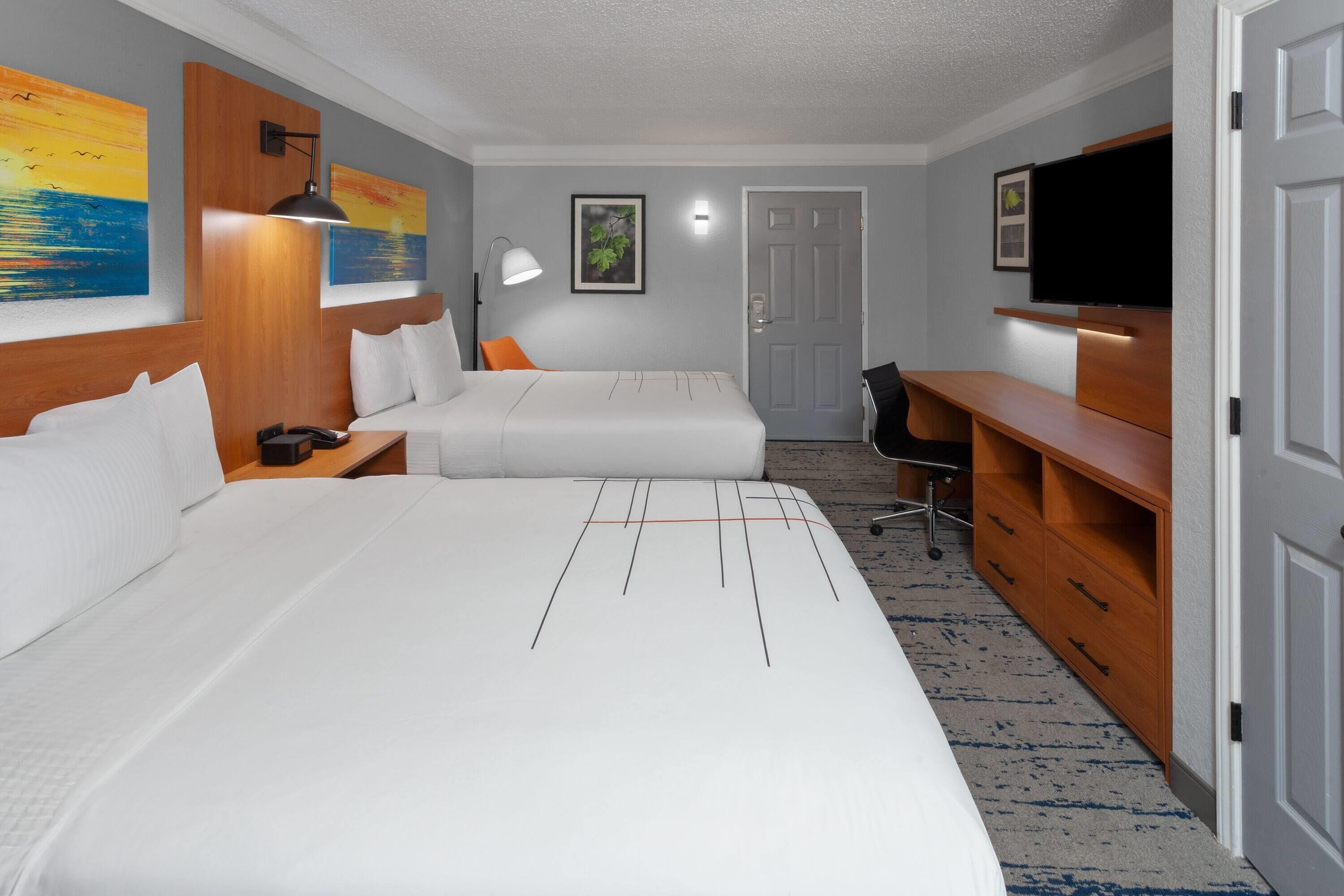 A hotel room with two beds, a desk, a chair, a TV, and artwork on the walls.