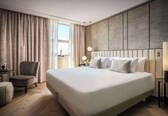A modern hotel room with a large bed, a seating area with a chair, a small table, and a view of a cityscape through the window.