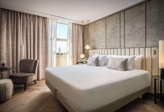 A modern hotel room with a large bed, a seating area with a chair, a small table, and a view of a cityscape through the window.