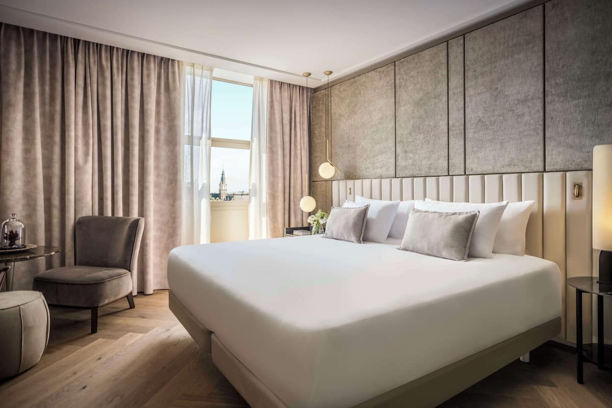 A modern hotel room with a large bed, a seating area with a chair, a small table, and a view of a cityscape through the window.