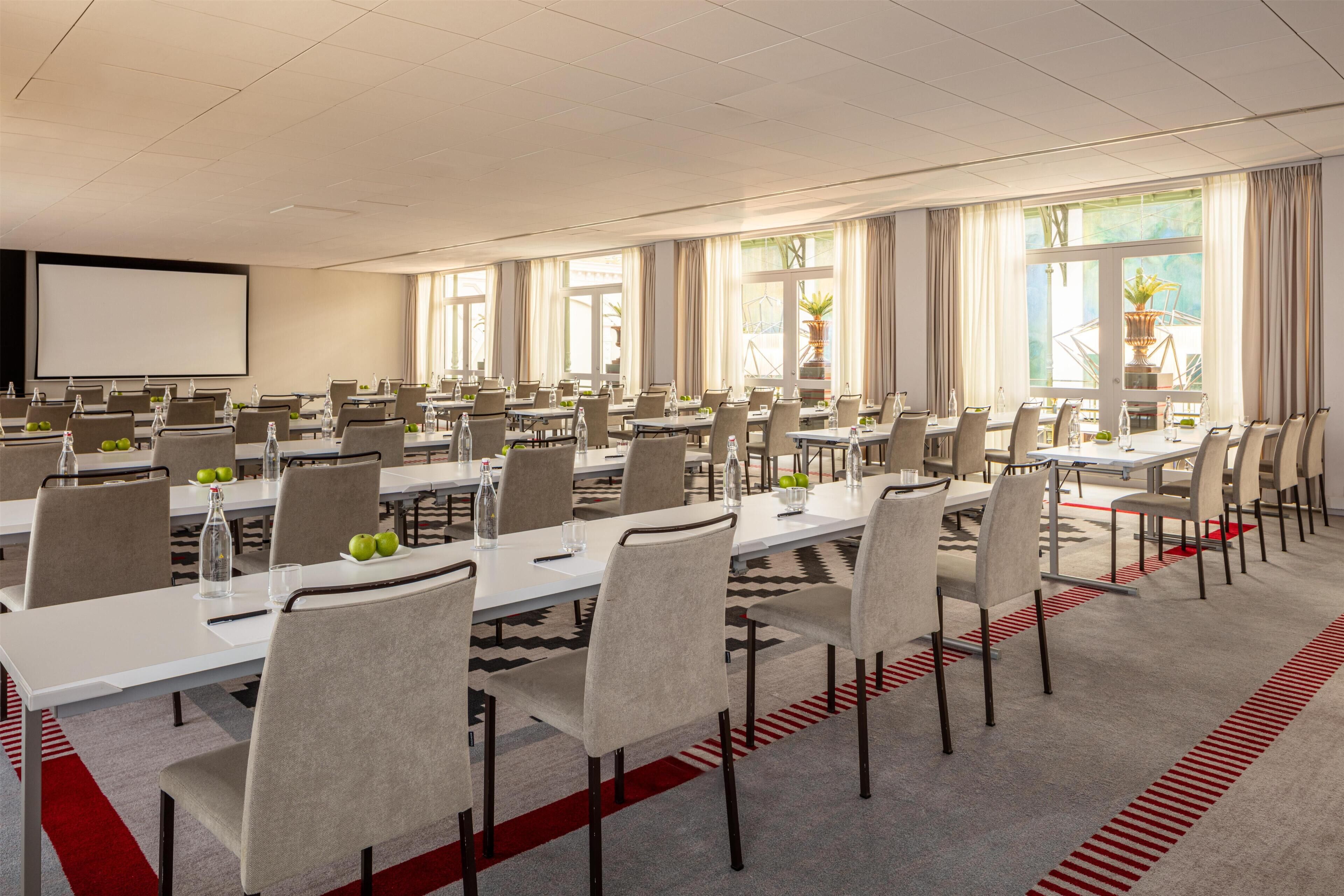 Un salón de conferencias con varias mesas y sillas, una pantalla de proyector y ventanas grandes con cortinas.