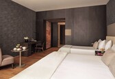 A modern hotel room with a large bed, a desk with a chair, a small table with a vase of flowers, and a large window.