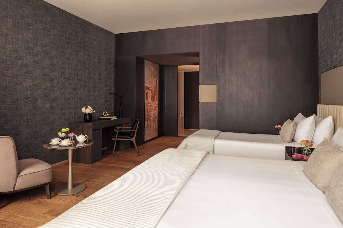 A modern hotel room with a large bed, a desk with a chair, a small table with a vase of flowers, and a large window.