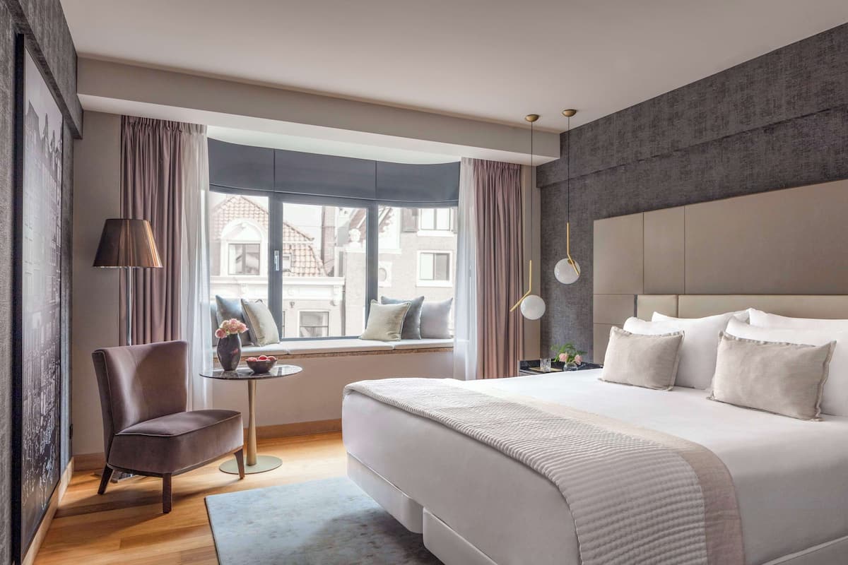 A modern hotel room with a large bed, a seating area with a chair and table, a view of buildings through the window, and a textured wall.