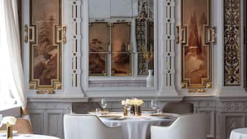 Un comedor con una mesa dispuesta para cuatro personas, rodeada de sillas blancas, con un ventanal decorativo y pinturas en la pared.