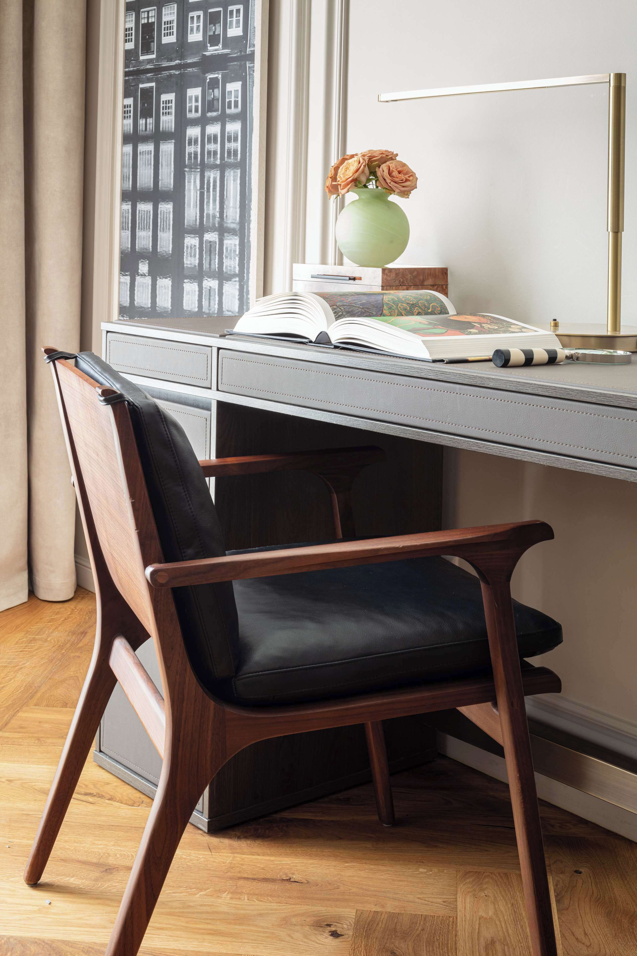 Une chaise en bois avec un coussin noir, placée devant un bureau près d’une fenêtre, avec un vase de fleurs et une lampe.