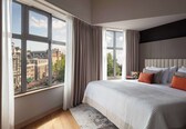 A modern bedroom with a large window, a bed with multiple pillows, and a view of buildings outside.