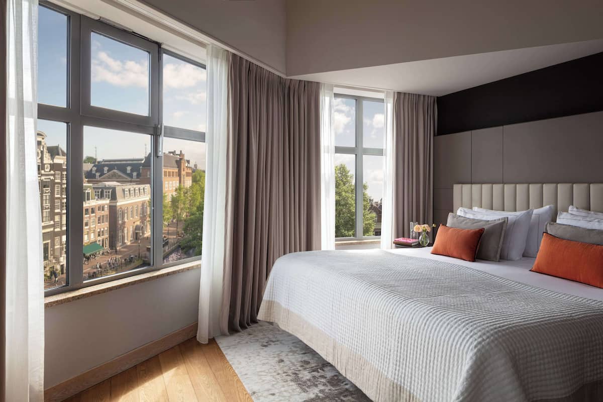 A modern bedroom with a large window, a bed with multiple pillows, and a view of buildings outside.