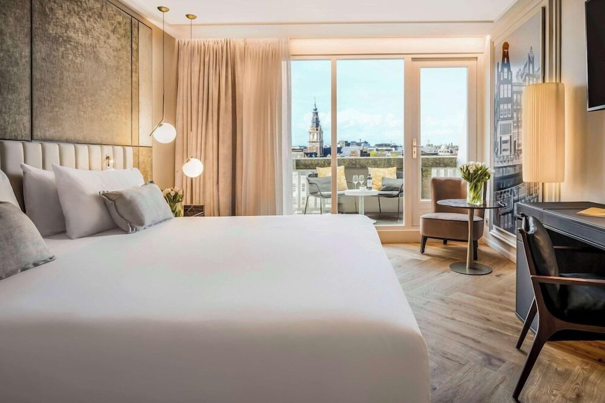 Habitación de hotel con una cama grande, un escritorio, una silla, un televisor y vista a la ciudad.