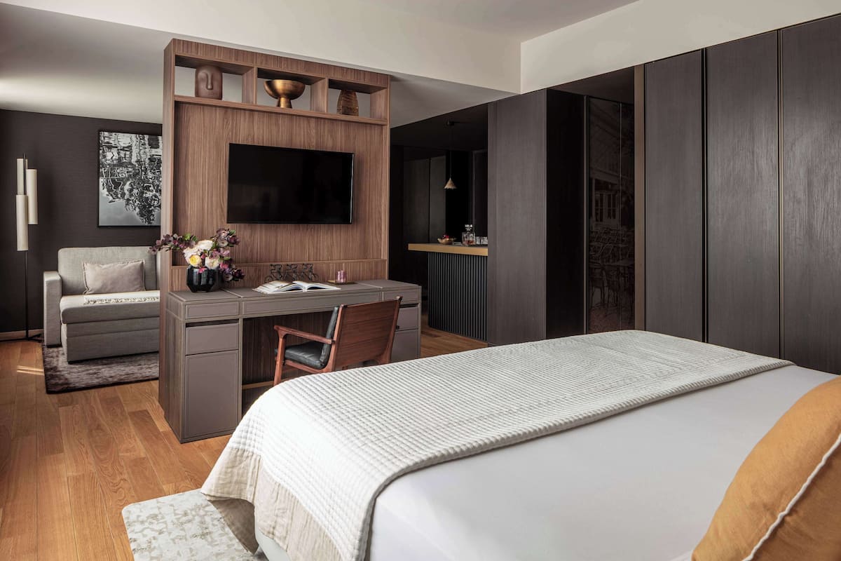 A modern hotel room with a large bed, a desk with a chair, a TV mounted on a wooden panel, and a sofa.