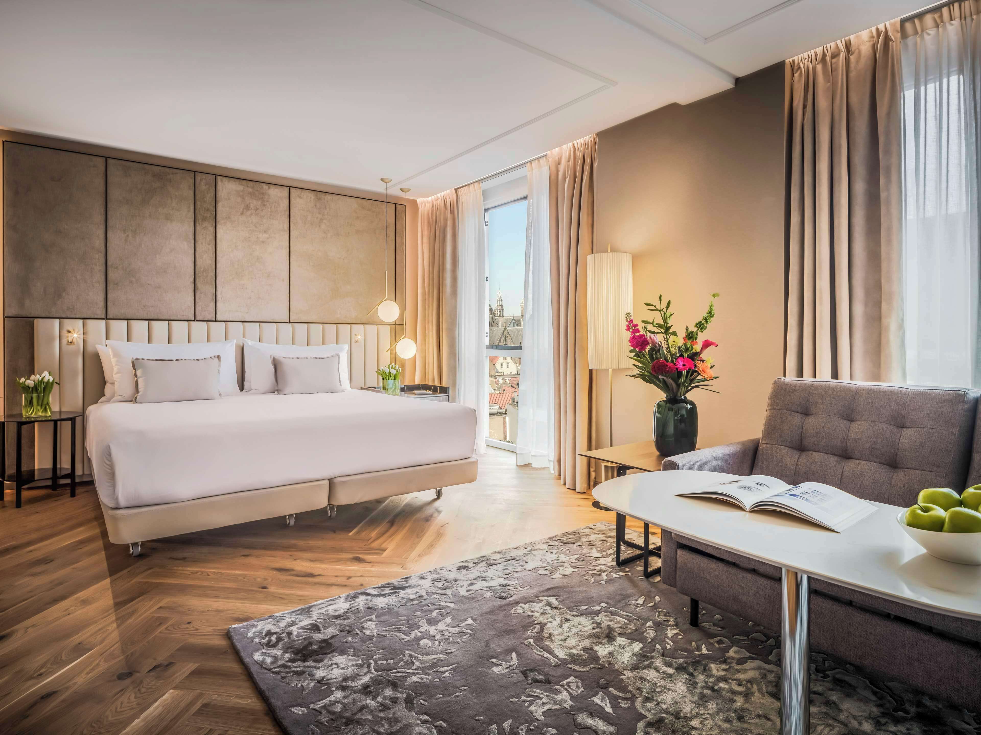 Una habitación de hotel moderna con una cama grande, un sofá, una mesita con un libro y manzanas, y un jarrón de flores.
