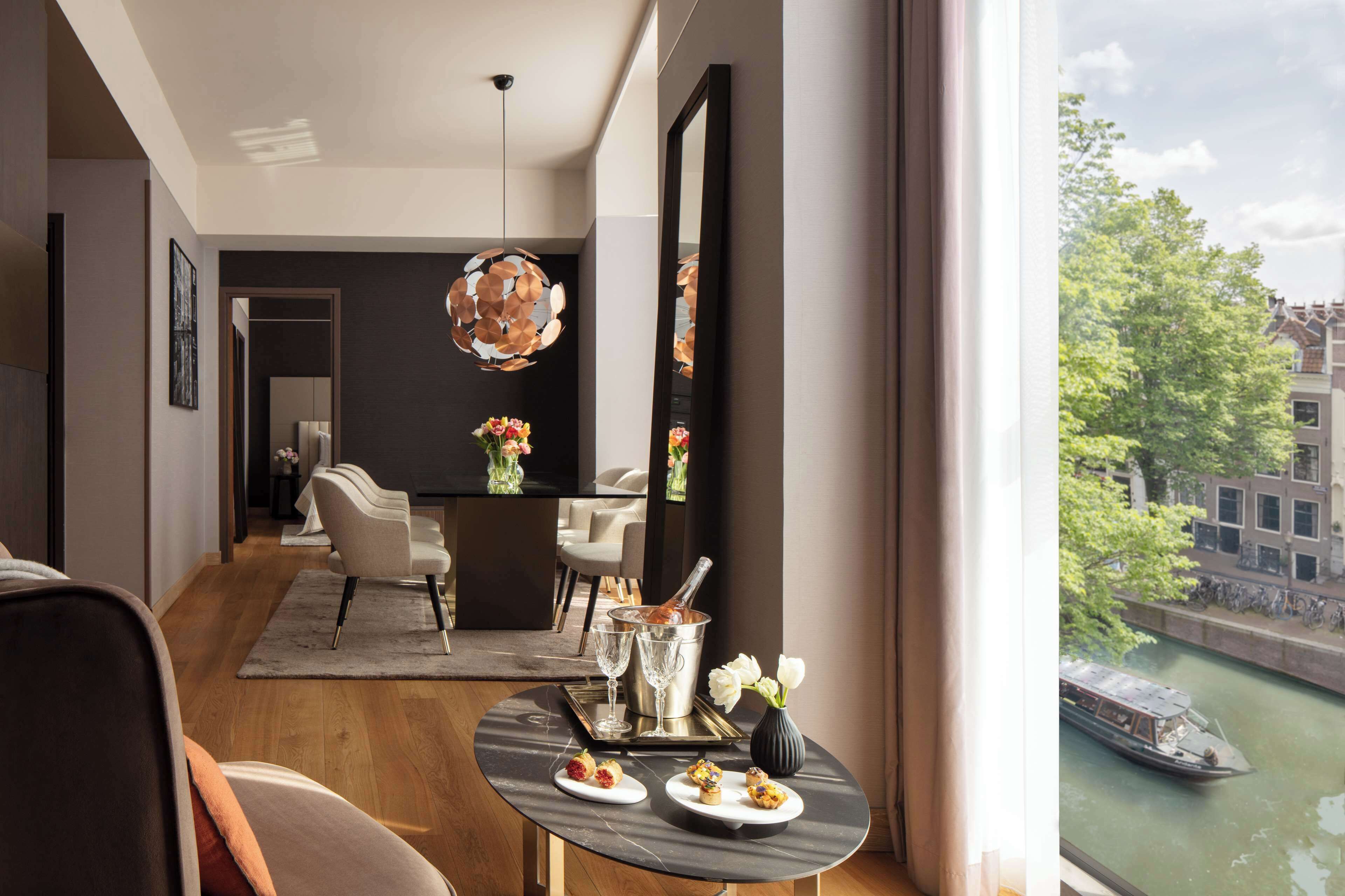 A modern hotel room with a dining area, a view of a canal, and a large mirror.