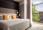 A modern bedroom with a large bed, a nightstand, a vase with flowers, and a view of the city through a large window.