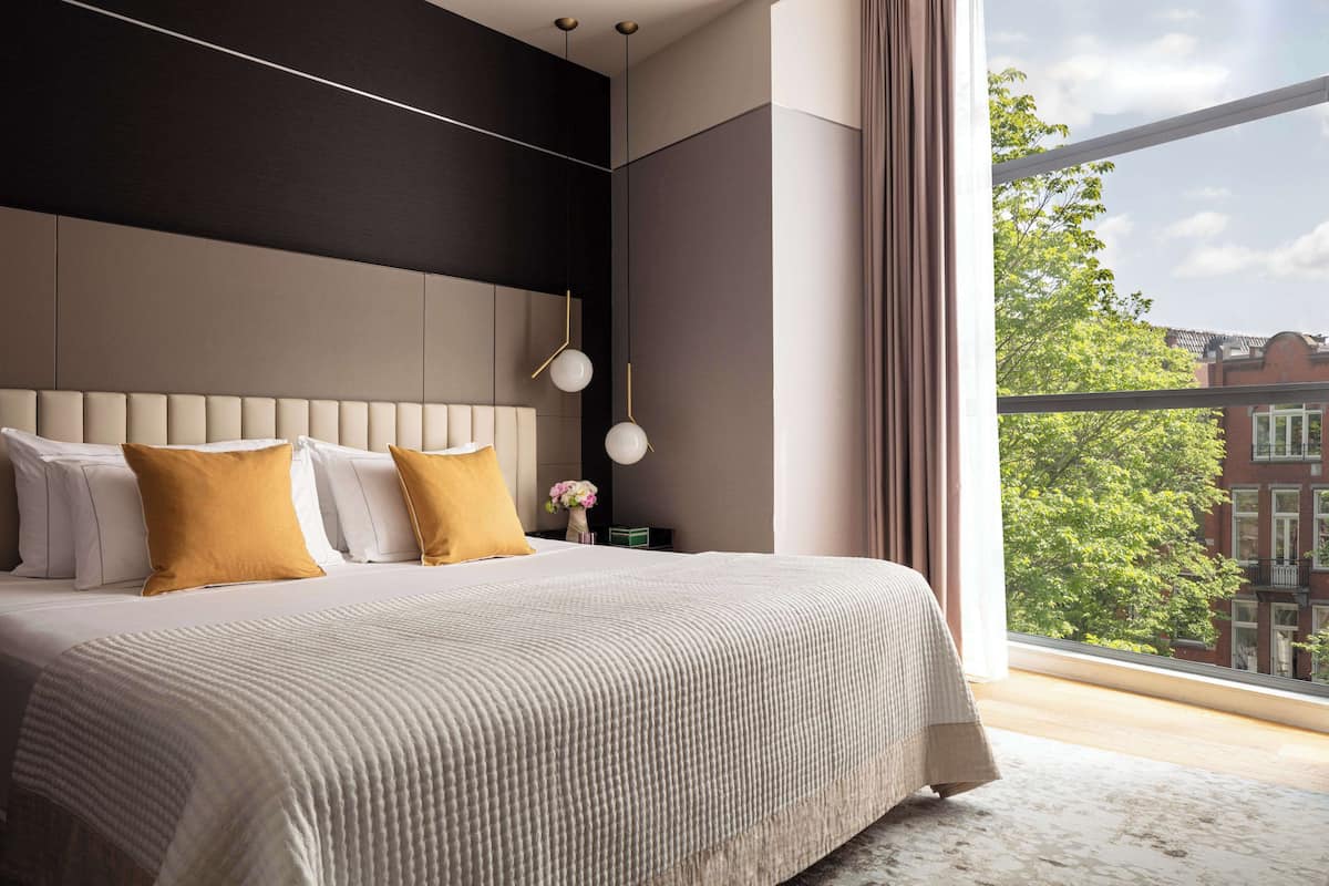 A modern bedroom with a large bed, a nightstand, a vase with flowers, and a view of the city through a large window.