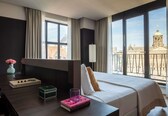 A hotel room with a large window, a bed, a bedside table with a book and a vase, and a view of a building with a dome.