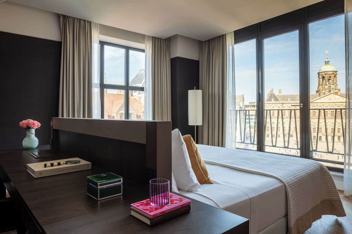 A hotel room with a large window, a bed, a bedside table with a book and a vase, and a view of a building with a dome.