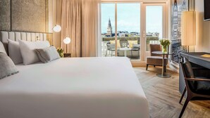 Une chambre d’hôtel avec un grand lit, un bureau, une chaise et une vue sur le paysage urbain grâce à de grandes fenêtres.