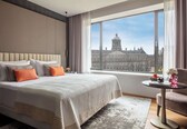 A modern hotel room with a large bed, a small round table, and a view of a historic building through a large window.